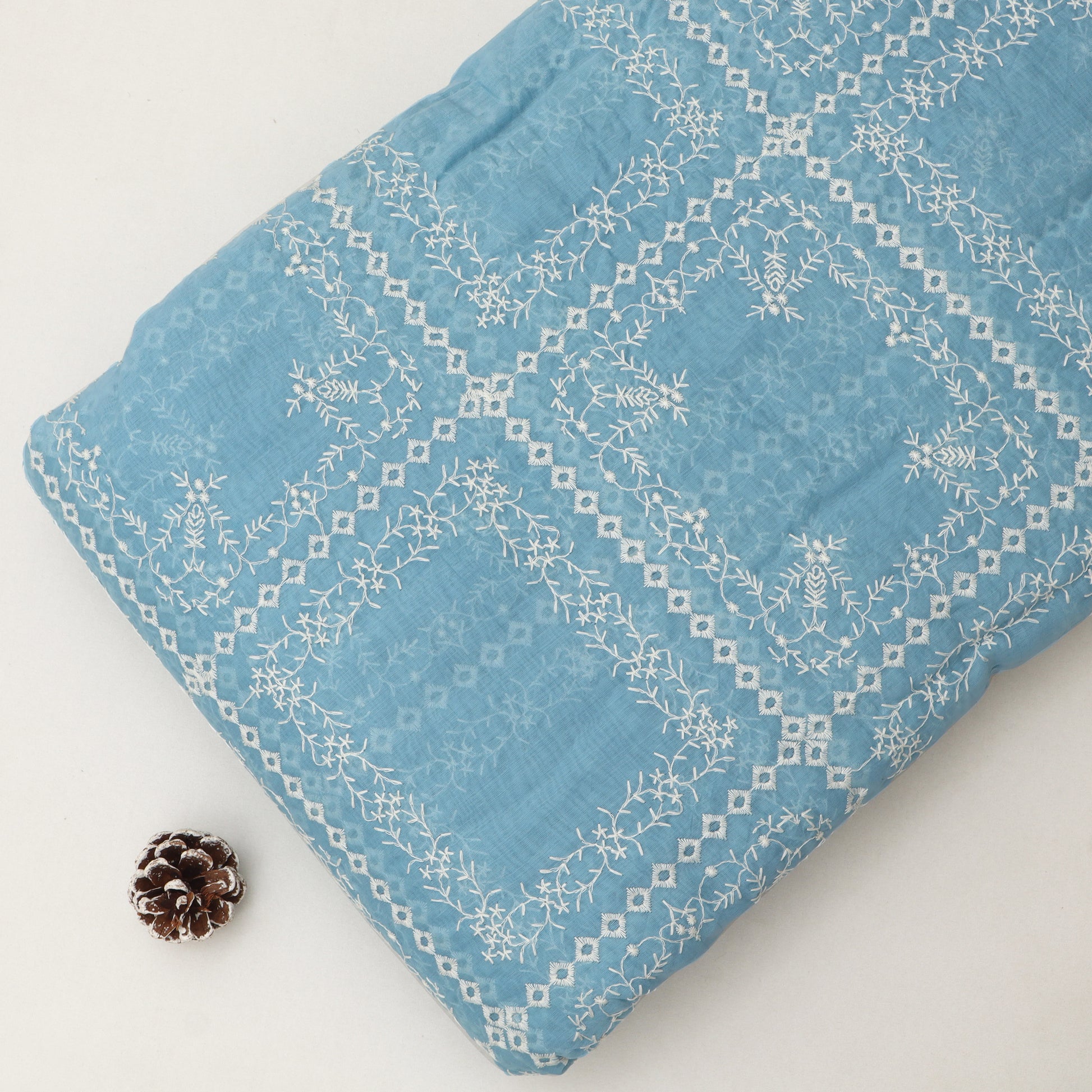 A folded light blue fabric item, likely a blanket or quilt, featuring white embroidered geometric and floral patterns, displayed on a light off-white background with a small pinecone prop.