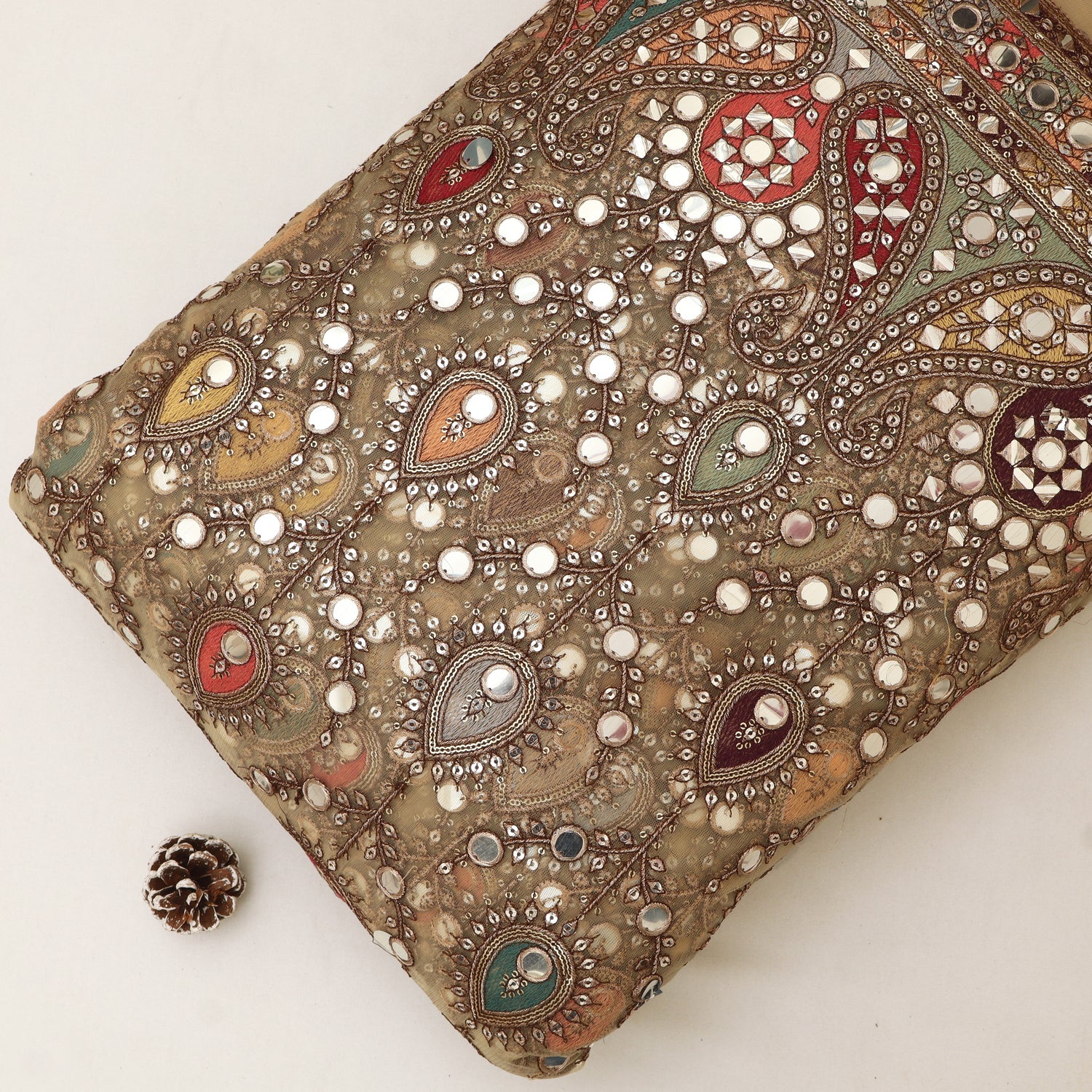A close-up of a beige and brown paisley fabric clutch or pouch heavily embellished with silver-toned mirror work and small white bead details, photographed on a plain light background with a small pinecone prop near the lower left.