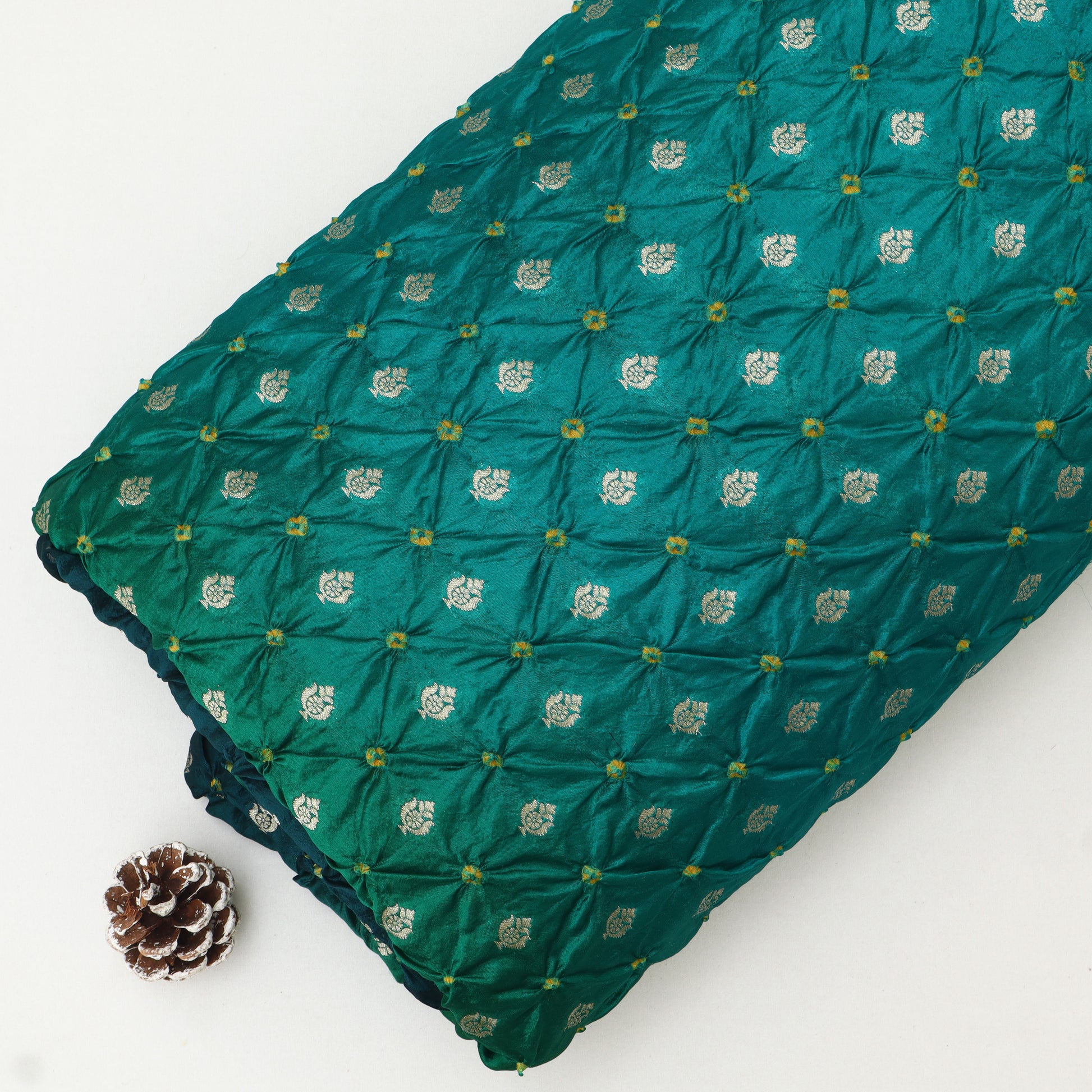 A folded green quilted textile with a repeating metallic gold floral pattern, displayed on a white background next to a small pinecone.