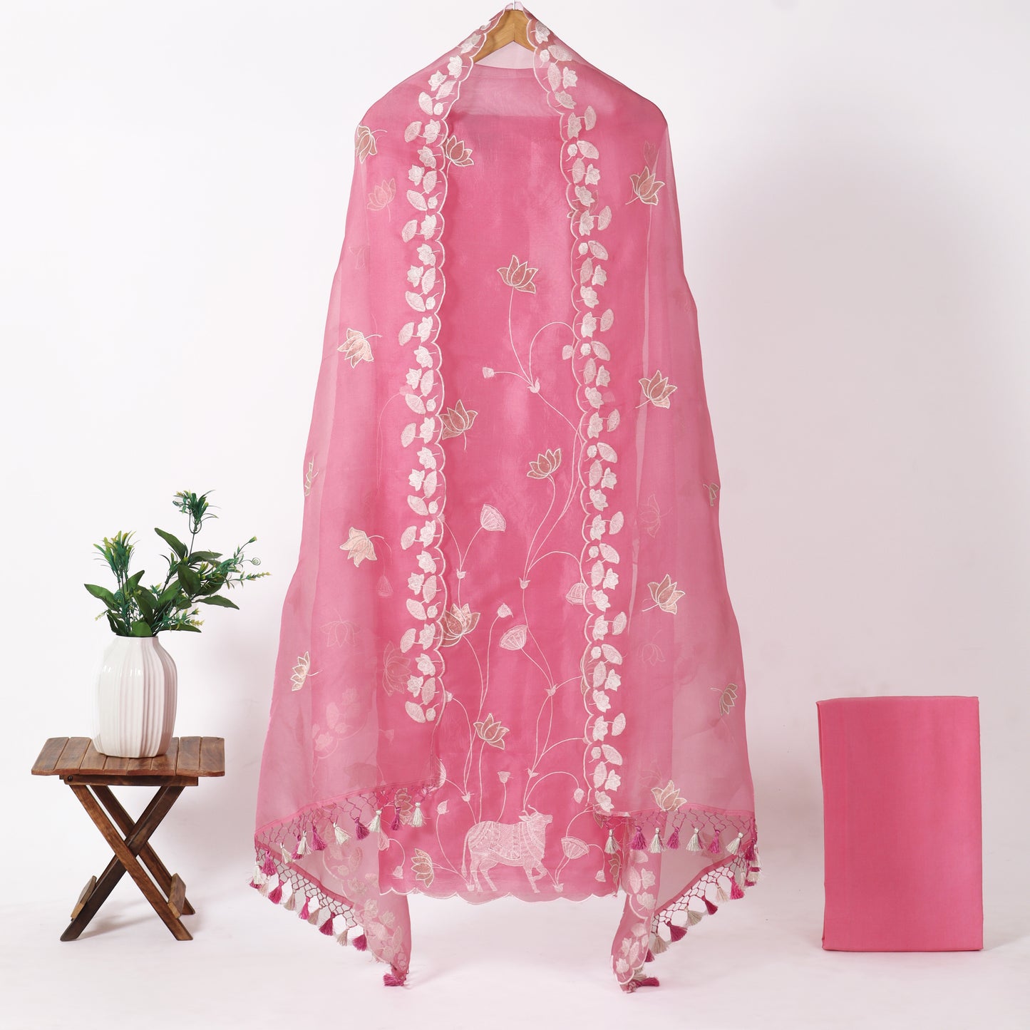 A pink sheer dupatta with white floral embroidery and tassel borders, hanging on a wooden hanger against a white background, with a small plant on a stool to the left and a folded matching pink fabric piece to the right.