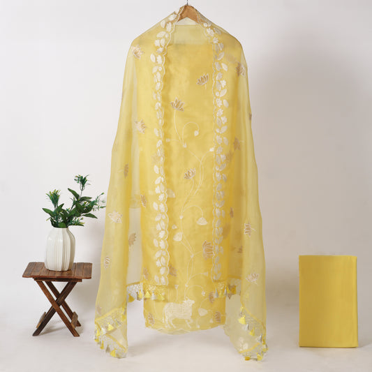 A sheer yellow shawl with white floral embroidery draped over a wooden hanger, styled on a light beige background with a small wooden stool holding a white vase with green leaves on the left, and a yellow rectangular box on the right.