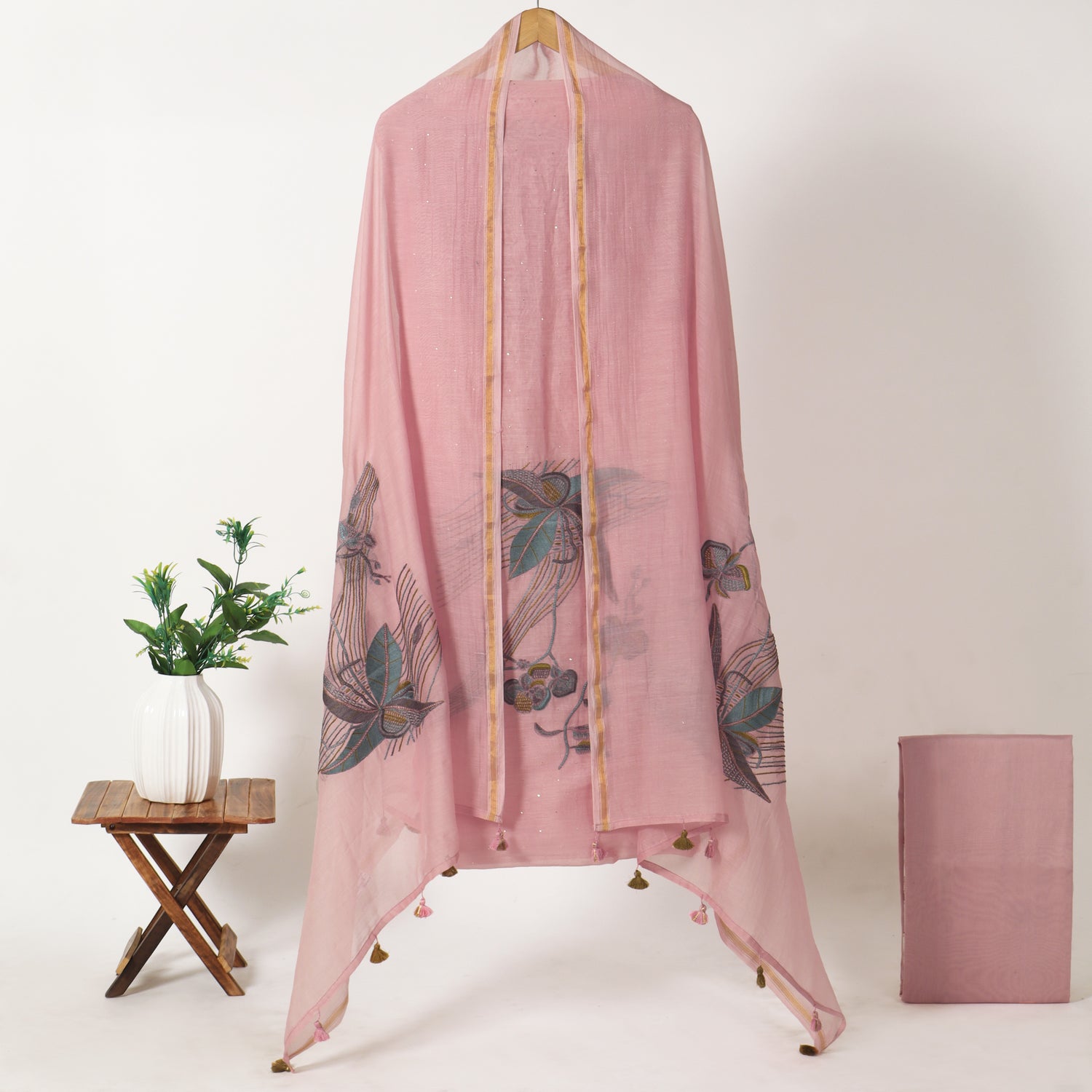 A light pink sheer scarf with butterfly and floral prints draped on a wooden hanger stand, with a matching folded scarf and a small potted plant beside it against a plain light background.