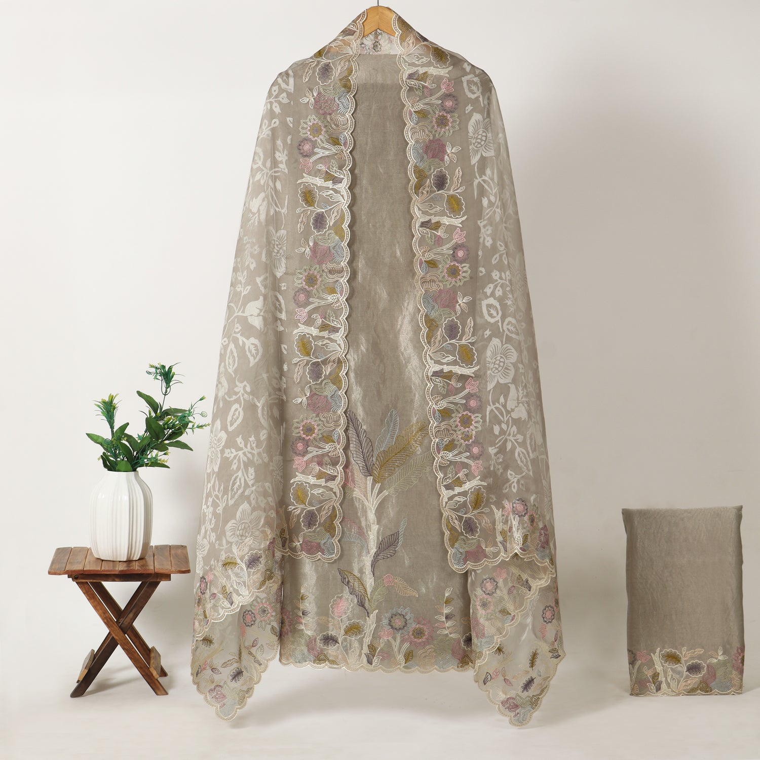 A sheer beige embroidered shawl or wrap with scalloped floral lace trim hangs on a wooden hanger against a light neutral background. A small wooden stool with a white vase of greenery sits to the left, and a folded matching fabric piece is placed to the right.