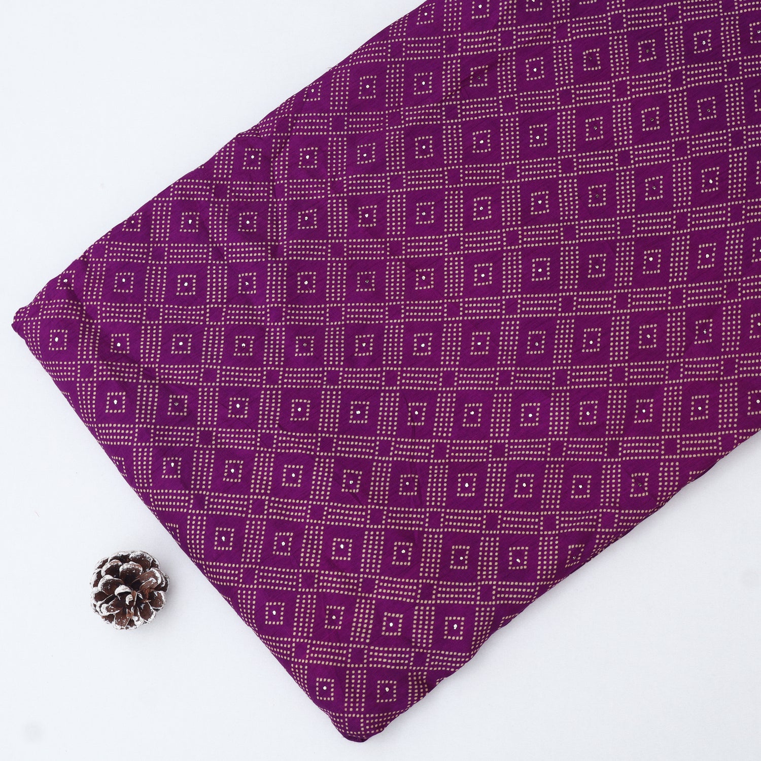 A folded piece of purple fabric with a repeating geometric diamond pattern, displayed on a white background next to a small metallic pinecone prop.