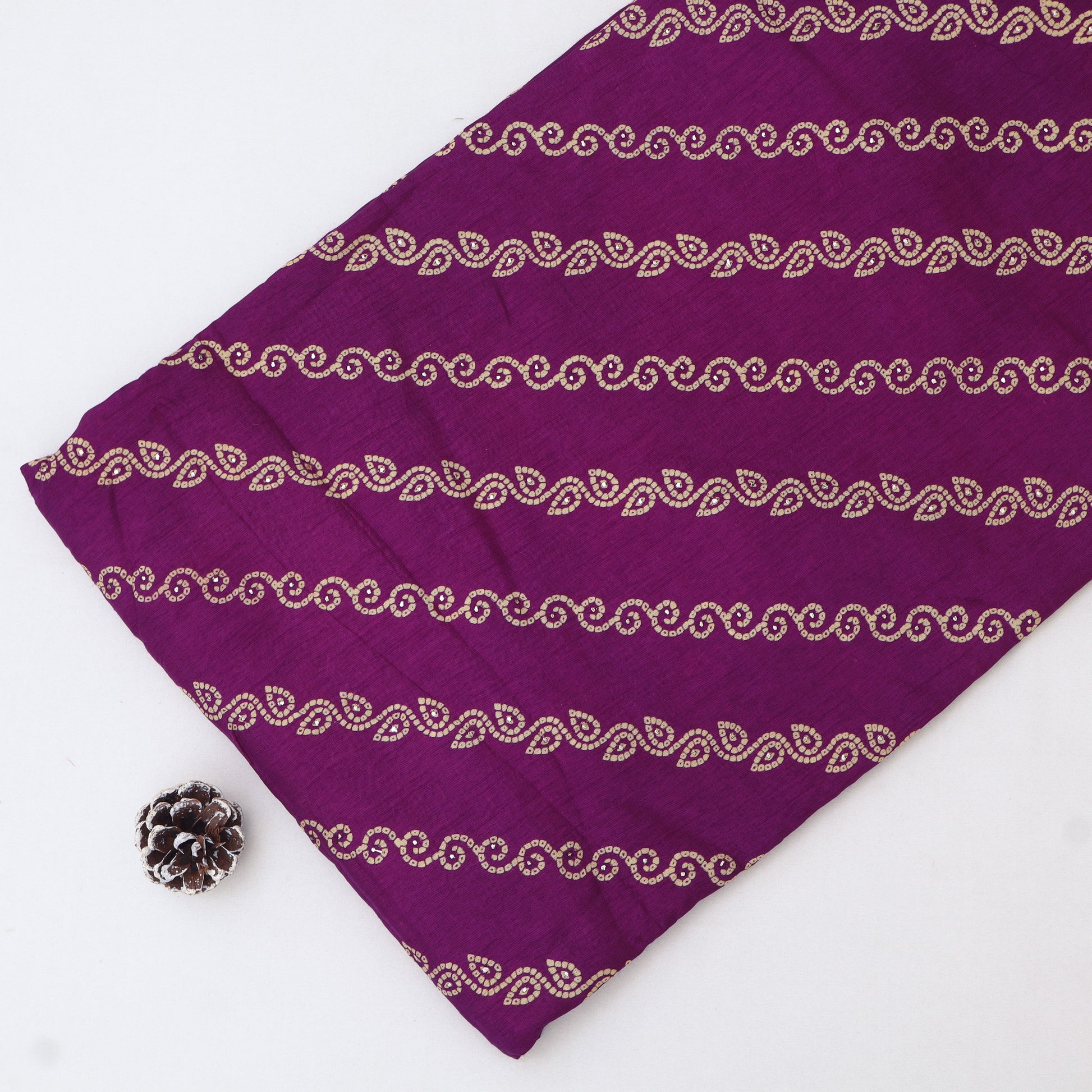 A close-up of a purple fabric with horizontal metallic gold paisley and wave patterns, laid flat on a white background with a small decorative pinecone prop in the lower-left corner.