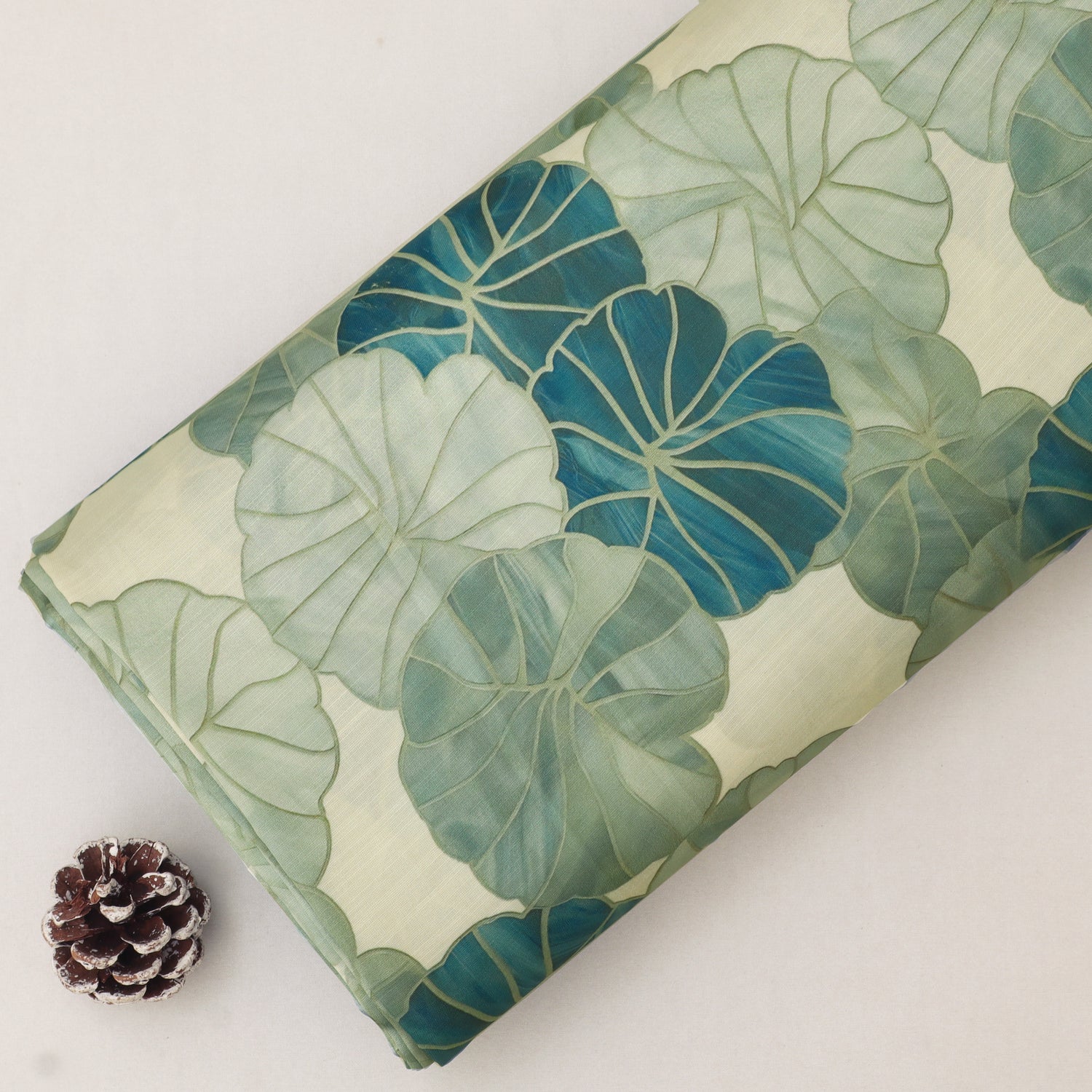 A folded fabric or paper item with a botanical leaf print in shades of teal, sage green, and cream, styled on a light background with a small pinecone prop.