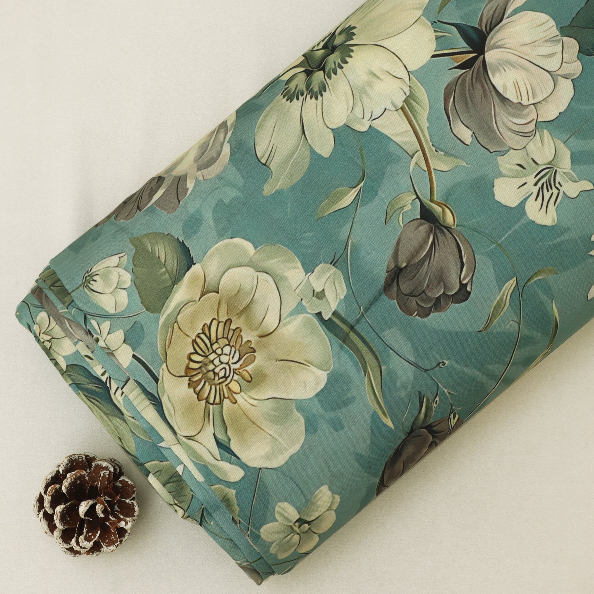 A folded piece of fabric with a floral print in teal, cream, and gray, styled with a small pinecone on a light beige background.