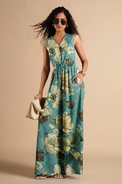 A woman modeling a sleeveless, V-neck maxi dress with a floral print in teal, green, and cream. She is wearing sunglasses, holding a small beige clutch, and standing against a solid beige studio background.