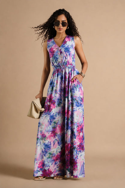 A woman modeling a sleeveless, V-neck maxi dress with a colorful watercolor floral print in shades of purple, pink, and blue. She is wearing dark sunglasses, a gold bracelet, and holding a small beige clutch bag against a solid beige studio background.