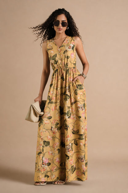 A woman modeling a sleeveless, V-neck maxi dress with a yellow floral and green vine print, holding a small beige clutch against a solid beige studio background.