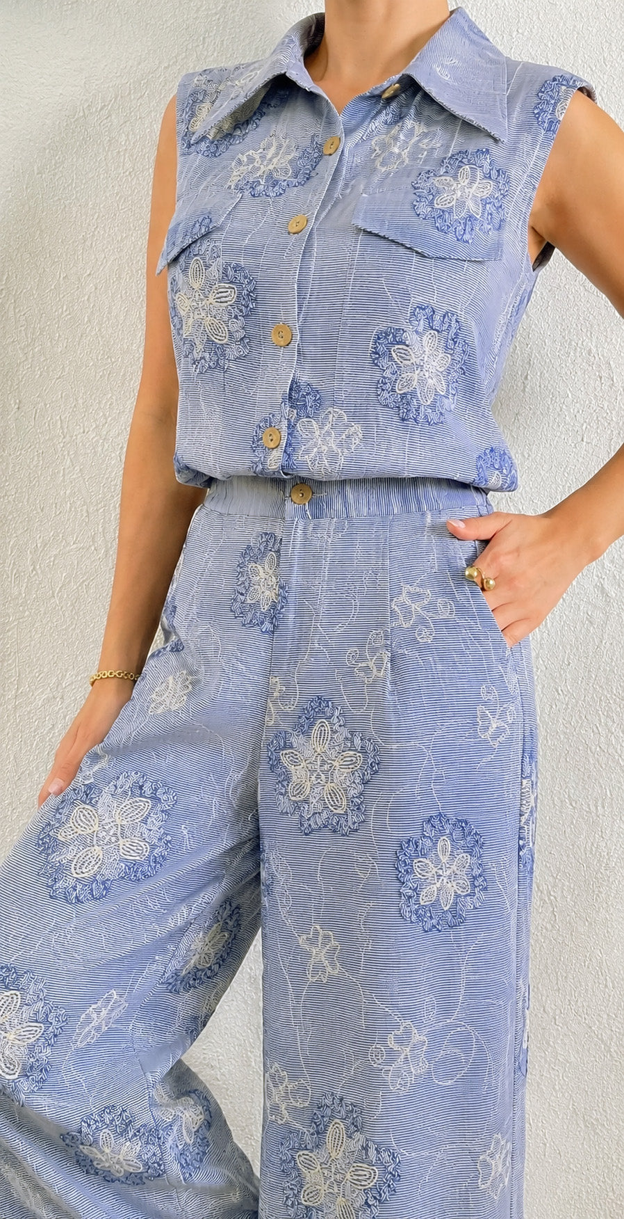 A sleeveless light blue collared button-front top and matching wide-leg pants set with a blue and white floral pattern, shown on a model against a textured off-white wall.