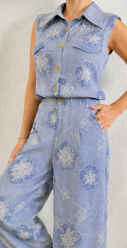 A sleeveless light blue collared button-front top and matching wide-leg pants set with a blue and white floral pattern, shown on a model against a textured off-white wall.