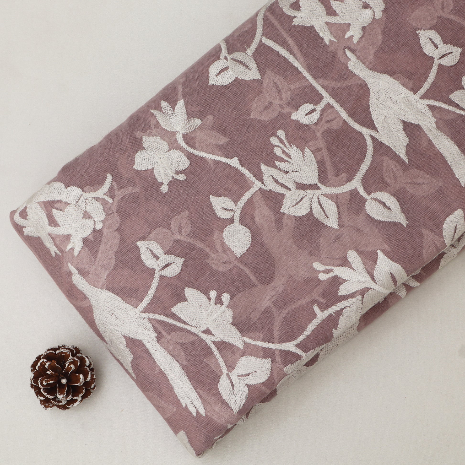 Lace fabric with white floral patterns on a mauve background, accompanied by a pine cone.