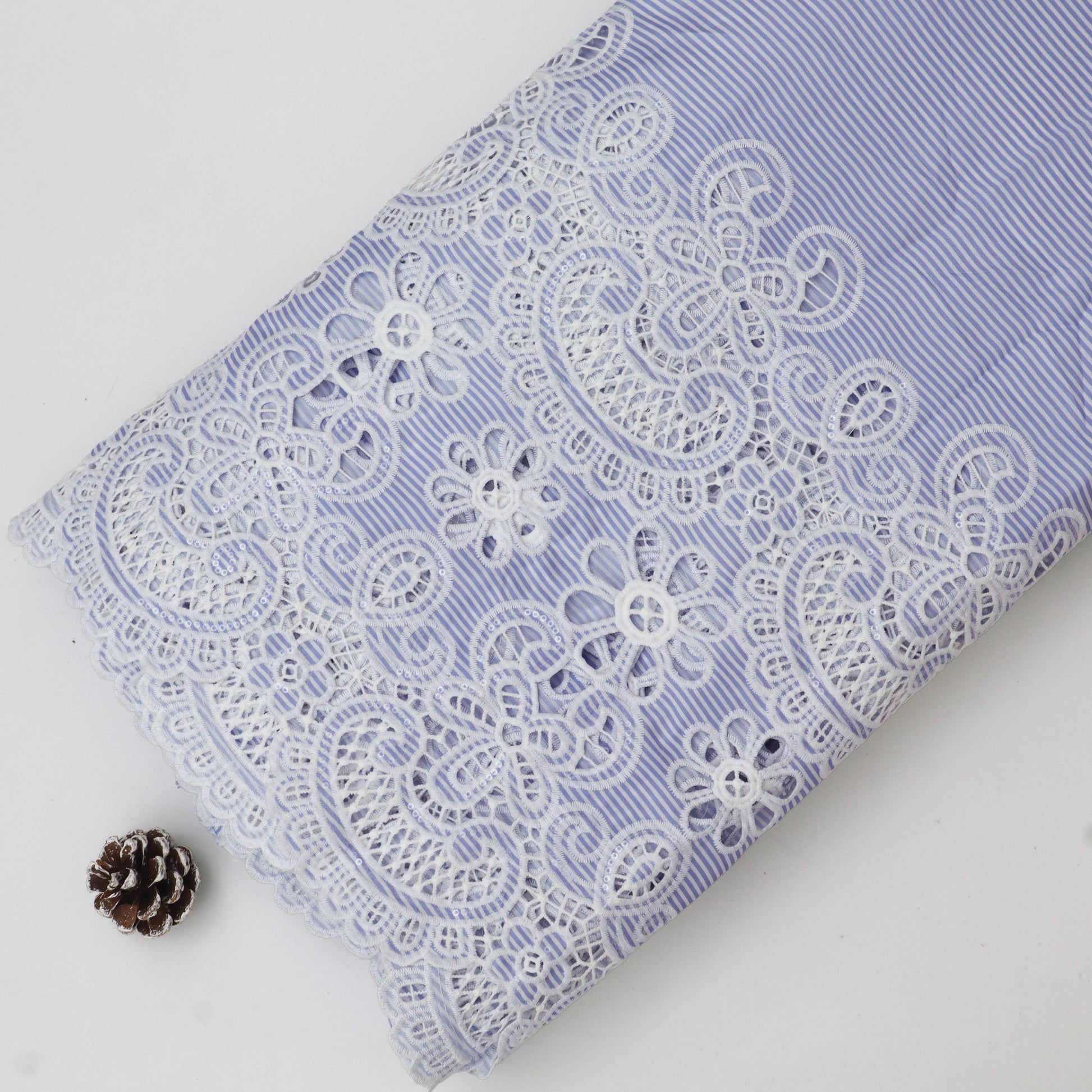 Lace fabric with white floral patterns on a light blue background, accompanied by a pine cone.