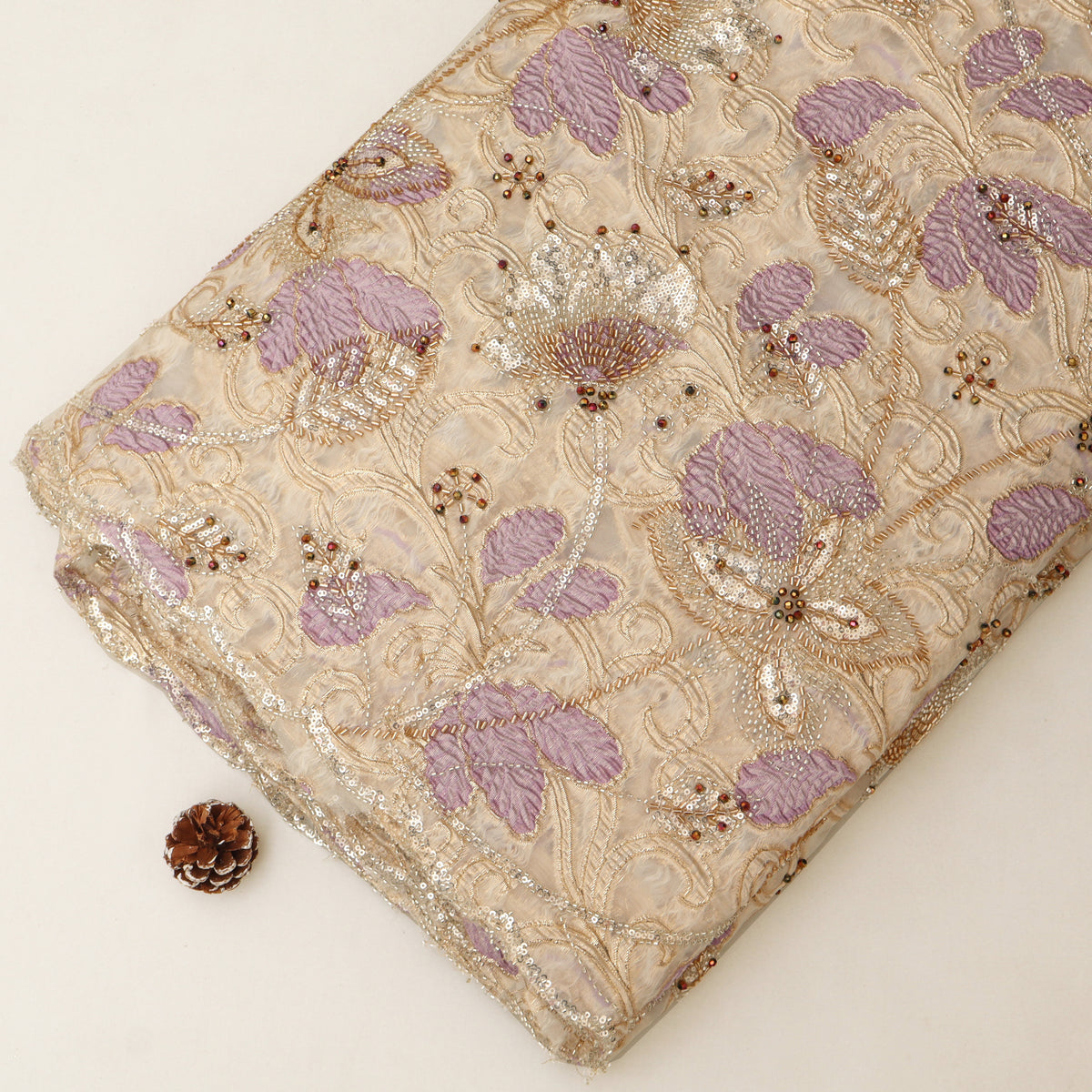 A folded piece of beige embroidered fabric with lavender floral motifs and metallic gold sequins, displayed on a light cream background next to a small pinecone.